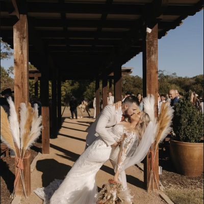 Bride and groom sharing first kiss under wooden pergola with pampas grass at Milagro Venue outdoor ceremony