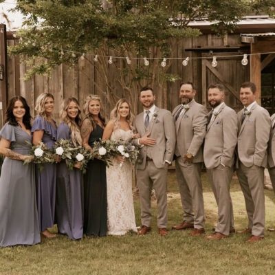 Wedding party group photo with slate blue bridesmaids in front of rustic barn at Milagro Venue Texas