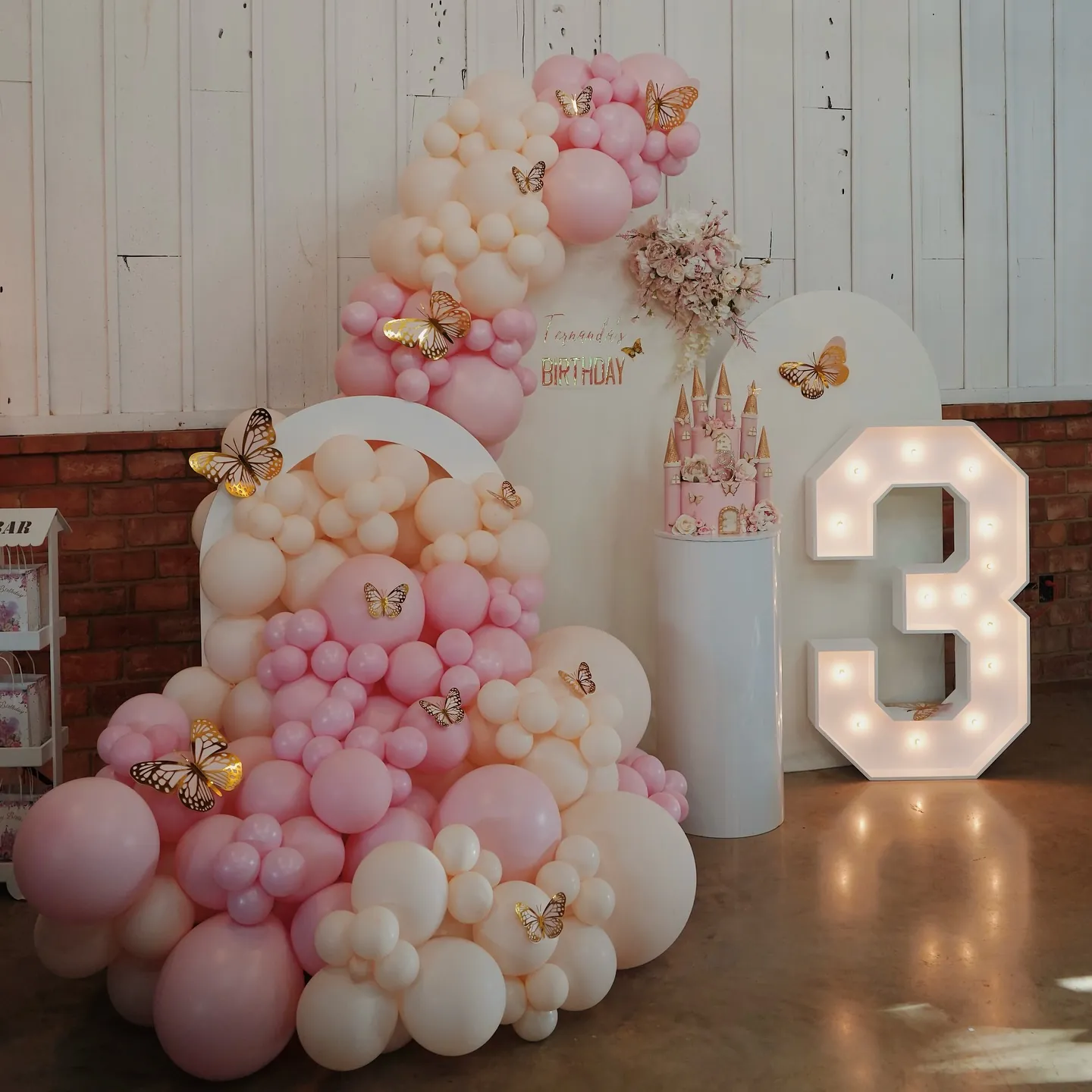 Pink balloon garland with gold butterflies, castle cake and lit number 3 marquee at a birthday party at Milagro Ranch